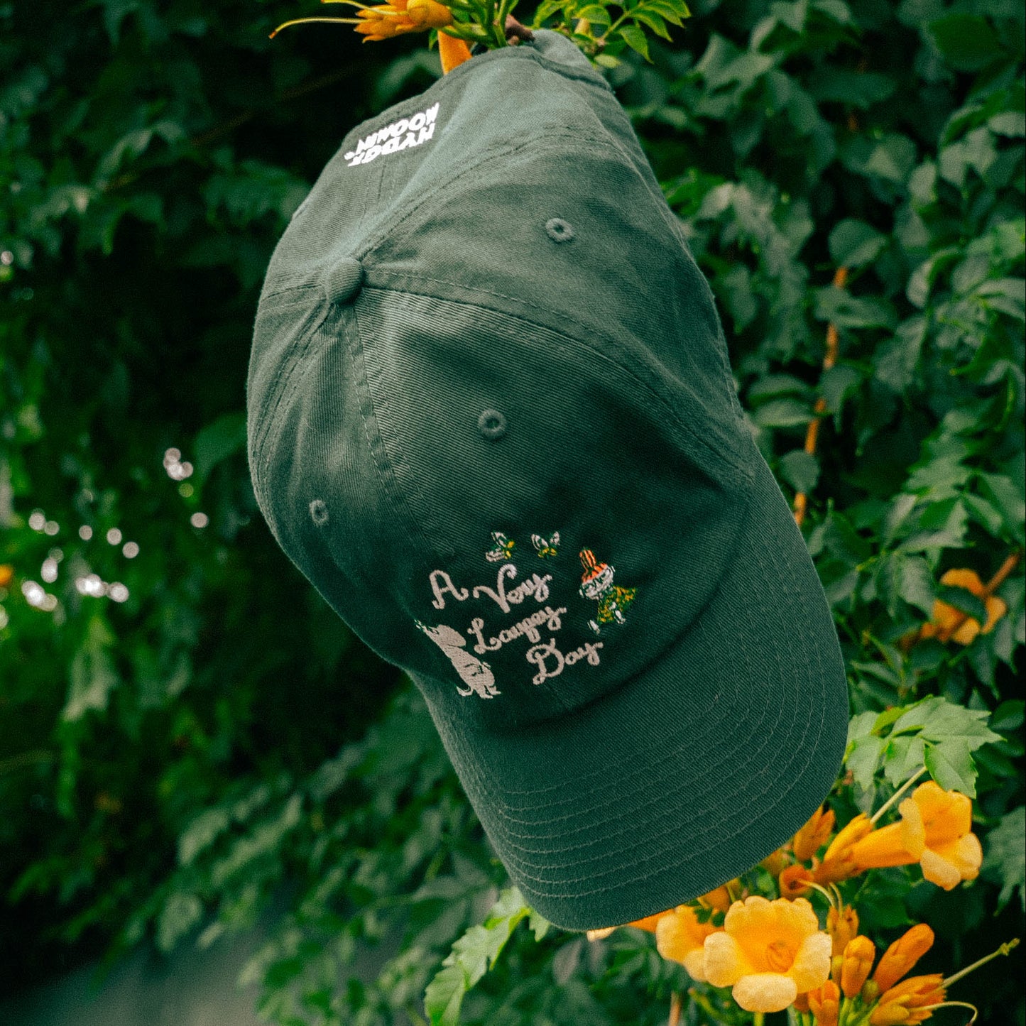 A dark green baseball cap with embroidered white text reading 'A very Laufey Day' and a small floral graphic, hanging in front of lush green foliage and orange flowers. 