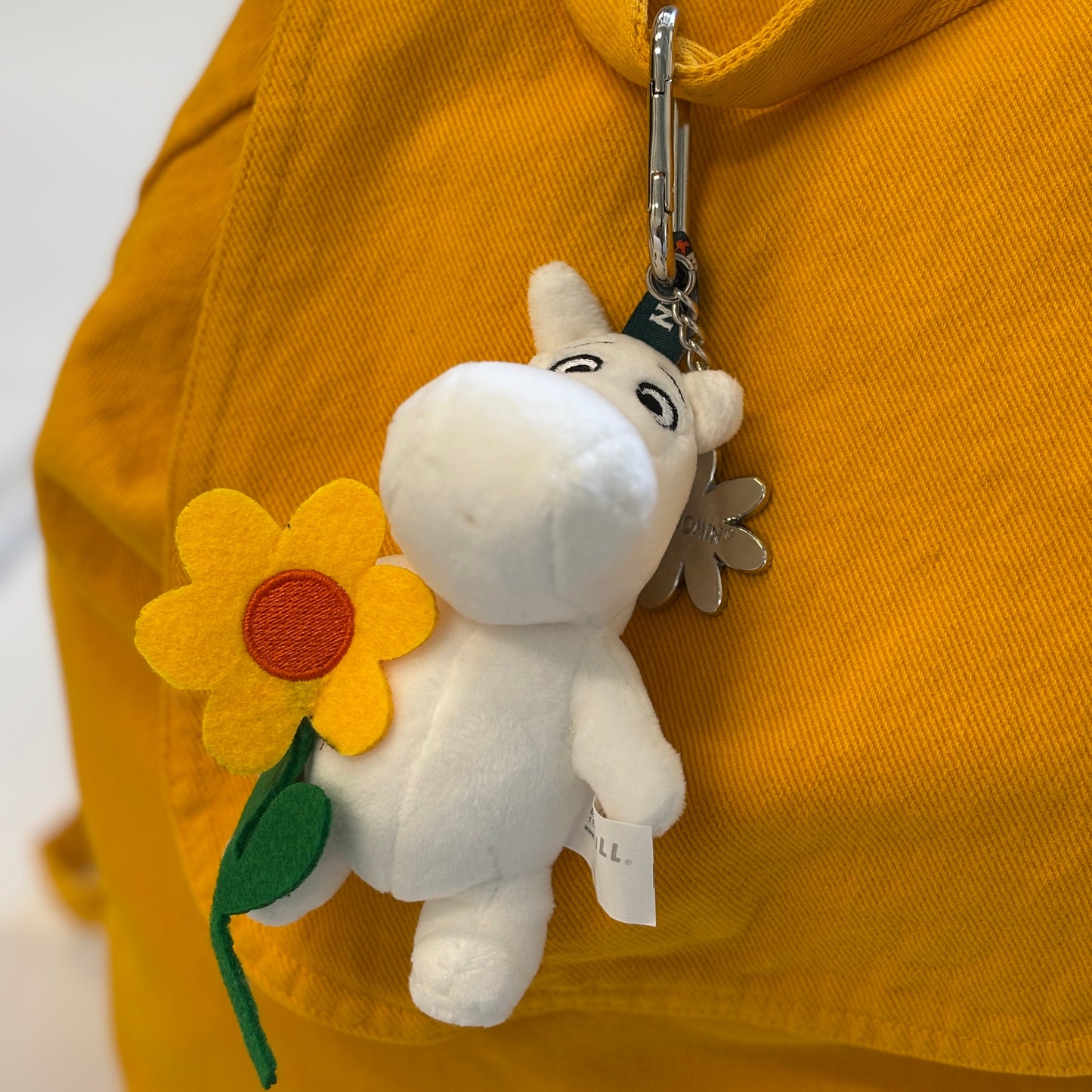 A white plush character keychain holding a yellow felt sunflower, attached to a silver carabiner and hanging on a bright yellow fabric bag.