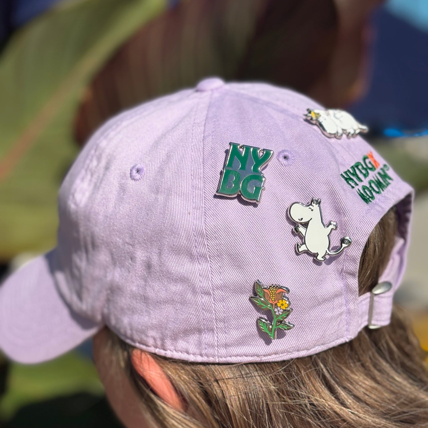 A close-up of a person wearing a light lavender baseball cap decorated with several enamel pins, including a green 'NY BG' pin, a white cartoon character pin, and a flower pin. The background is heavily blurred with green foliage and warm tones.