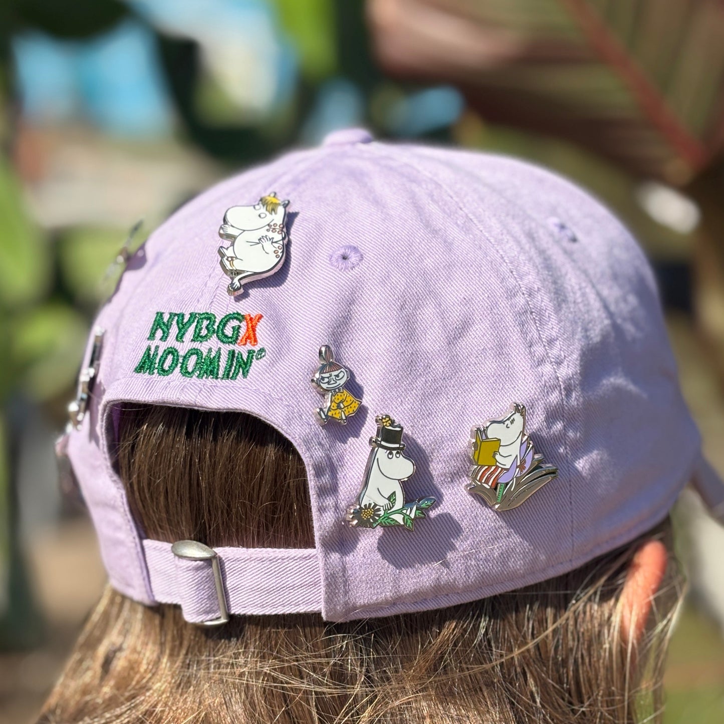 A lavender purple baseball cap with 'NYBG x MOOMIN' pins and Moomin character enamel pins, shown from the back on a person with long brown hair in an outdoor garden setting.