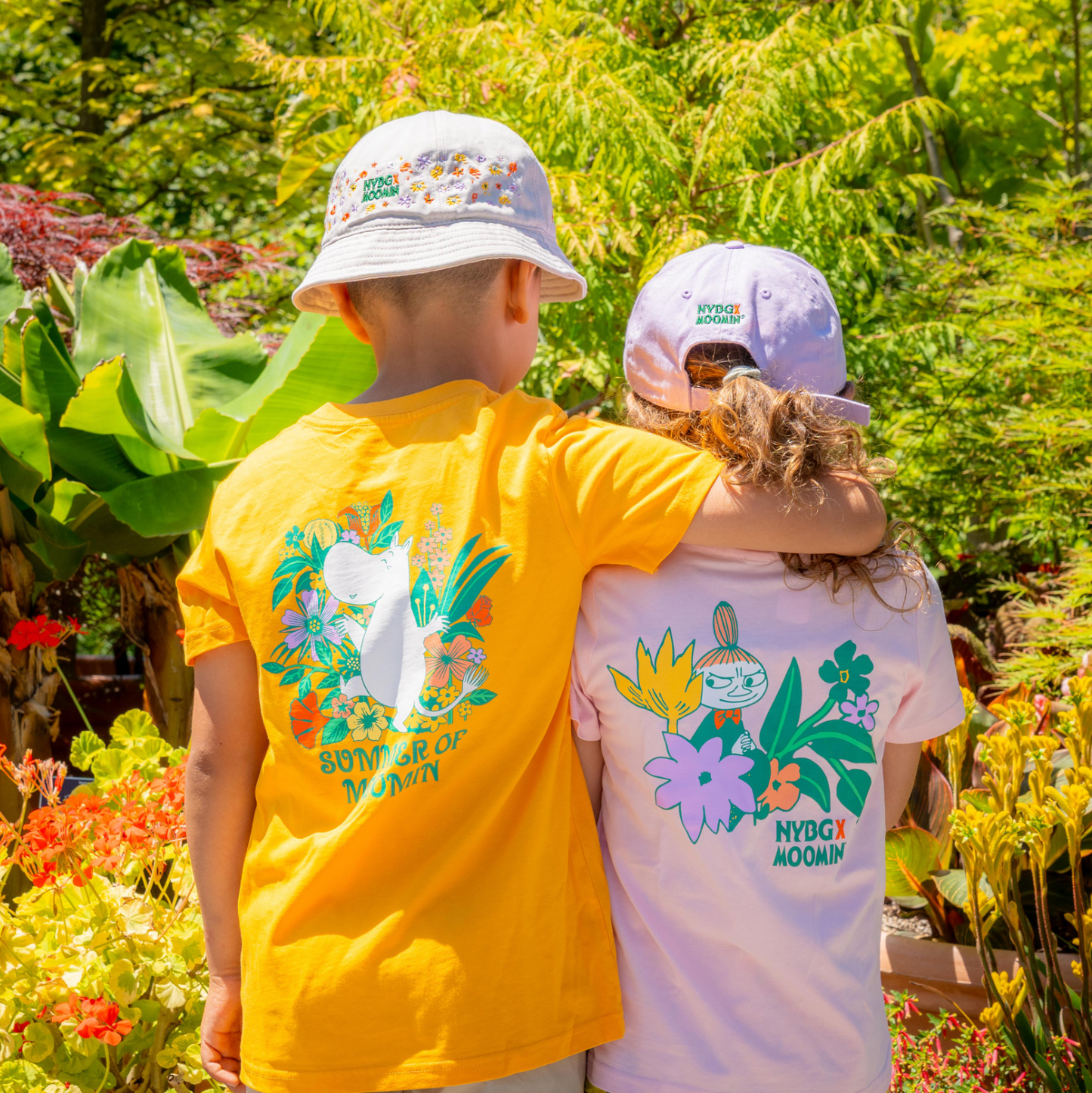 Two children standing outdoors in a garden, viewed from behind. The child on the left wears a bright yellow T-shirt with a large colorful floral bunny graphic and the words 'BUNN OF MARCH' on the back, along with a white bucket hat.